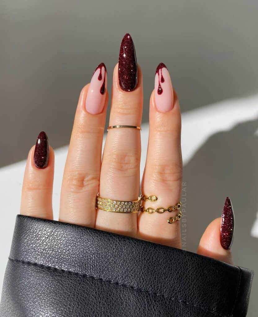 Hand with long almond shaped nails featuring deep burgundy glitter polish paired with soft nude pink accent nails and subtle drip style details, creating a bold February manicure with a moody, Valentine inspired feel.