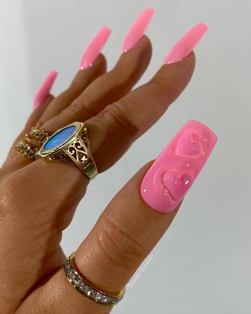 Hand with long square pink nails featuring a glossy finish and raised 3D heart accents on the accent nail, creating a bold Valentine’s inspired manicure against a neutral background.