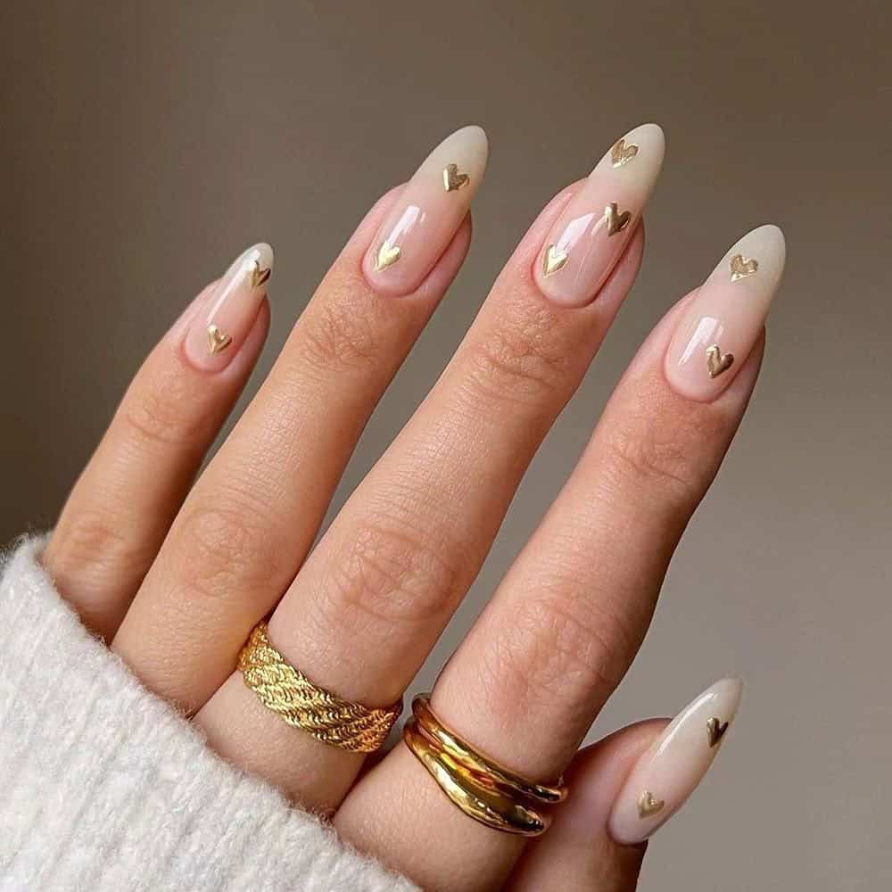 Hand with almond. shaped nude ombré nails featuring soft milky tips and tiny gold heart accents, creating an elegant February manicure with a subtle Valentine’s inspired look.
